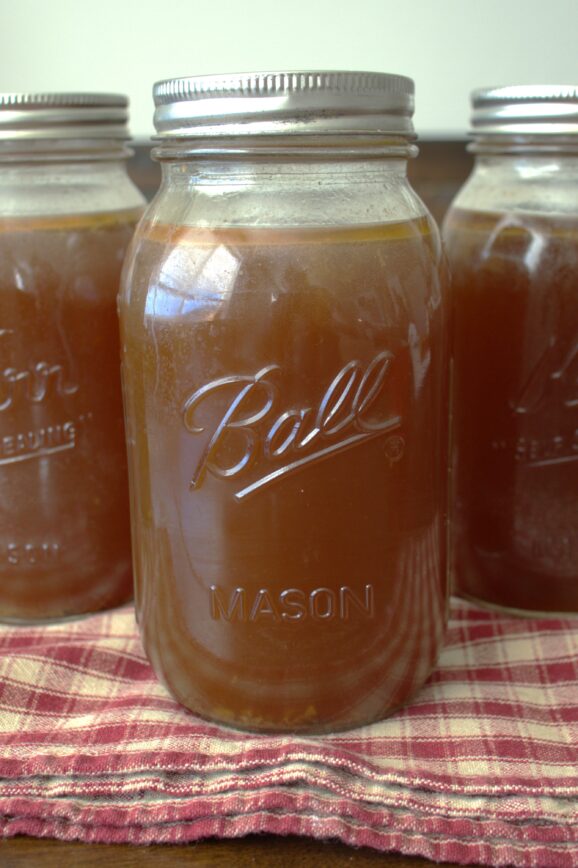 Three quart jars of dark chicken broth on a checkered red towel on top of a wooden table