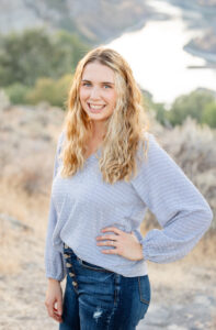 Girl standing in front of a river in a canyon with her and on her hip, smiling.