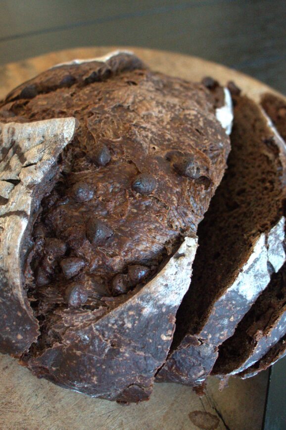 A sourdough double chocolate boule loaf on a round wooden cutting board with three slices cut off of the right side and a serrated knife.