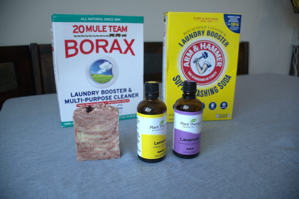 A box of borax, washing soda, a non-toxic soap bar, and lemon and lavender essential oils on a table with a blue table cloth.
